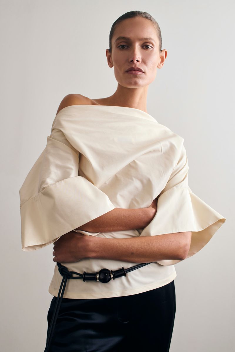 Fashion shot of a woman in an off-the-shoulder top, with a black belt, against a light grey background. The photograph is a studio shot.