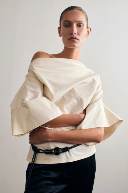 Fashion shot of a woman in an off-the-shoulder top, with a black belt, against a light grey background. The photograph is a studio shot.