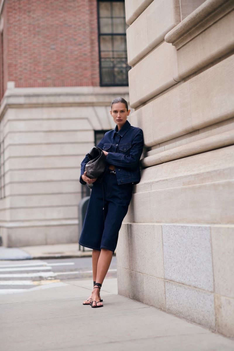 Model leaning against a stone building wearing a dark blue denim jacket and matching shorts, holding a dark leather bag.