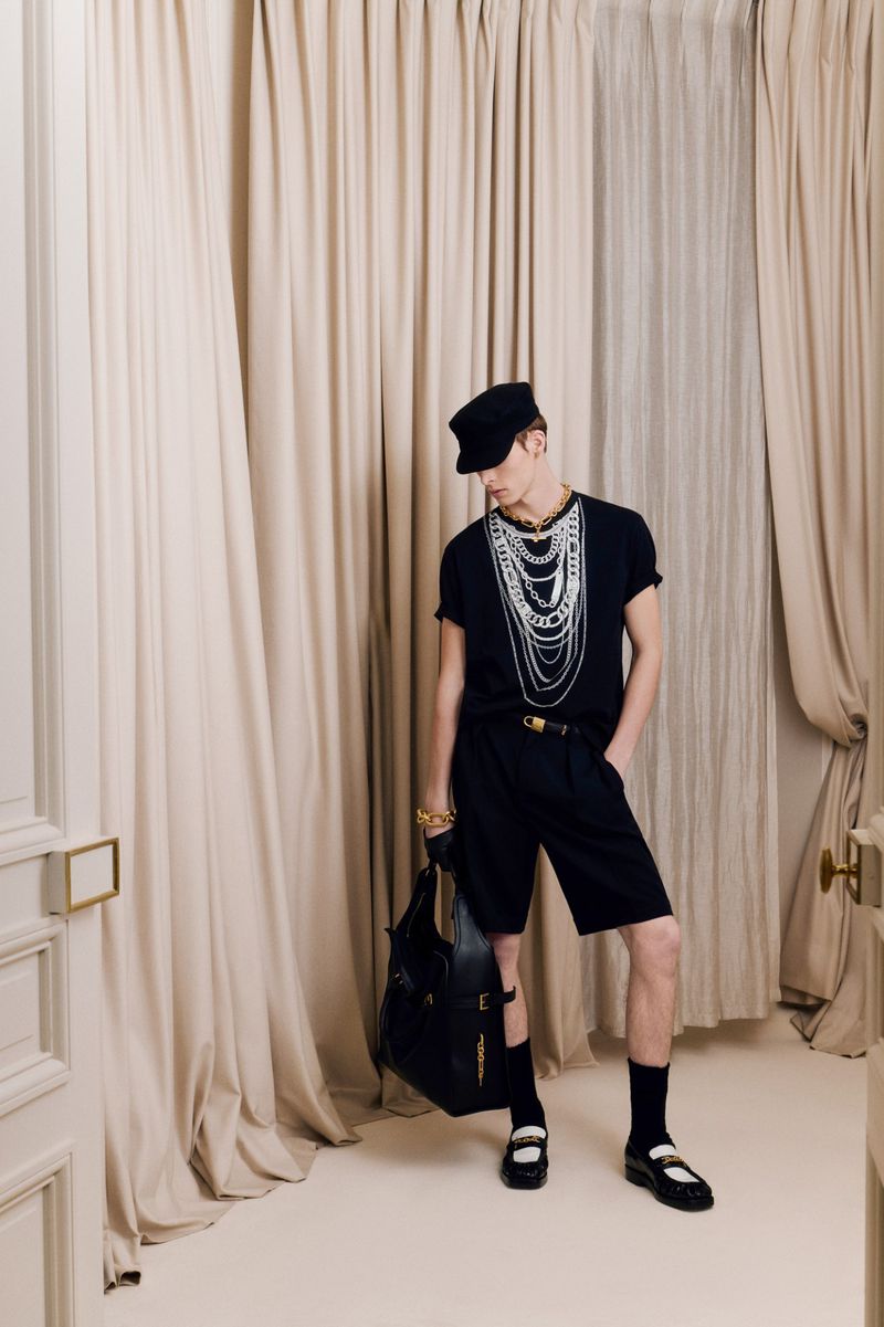 Fashionable adult male poses indoors wearing a black shirt, shorts, accessories, and black handbag, standing against cream-colored curtains.