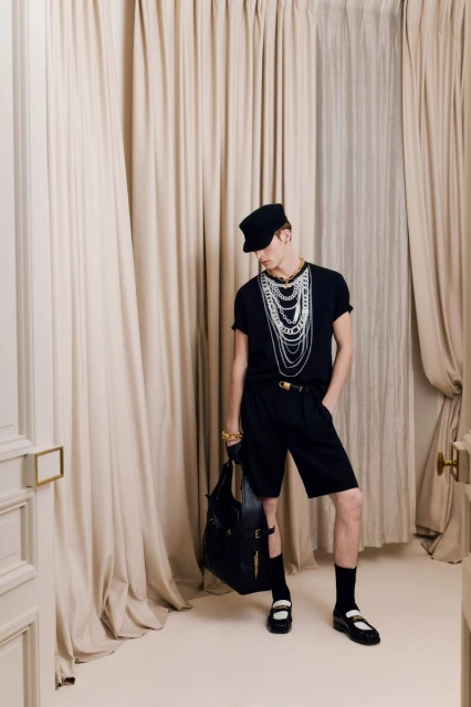 Fashionable adult male poses indoors wearing a black shirt, shorts, accessories, and black handbag, standing against cream-colored curtains.