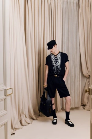 Fashionable adult male poses indoors wearing a black shirt, shorts, accessories, and black handbag, standing against cream-colored curtains.