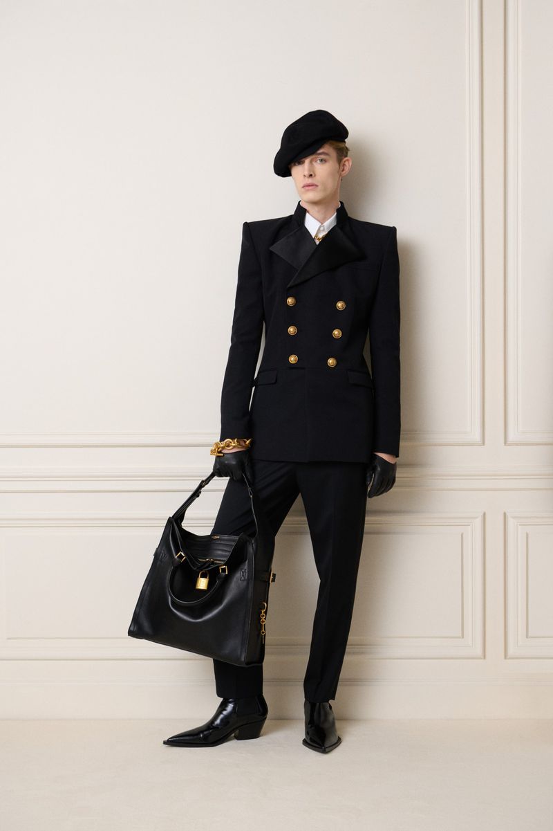Full-length shot of a person wearing a black double-breasted suit with gold buttons, black beret, and holding a black leather handbag.