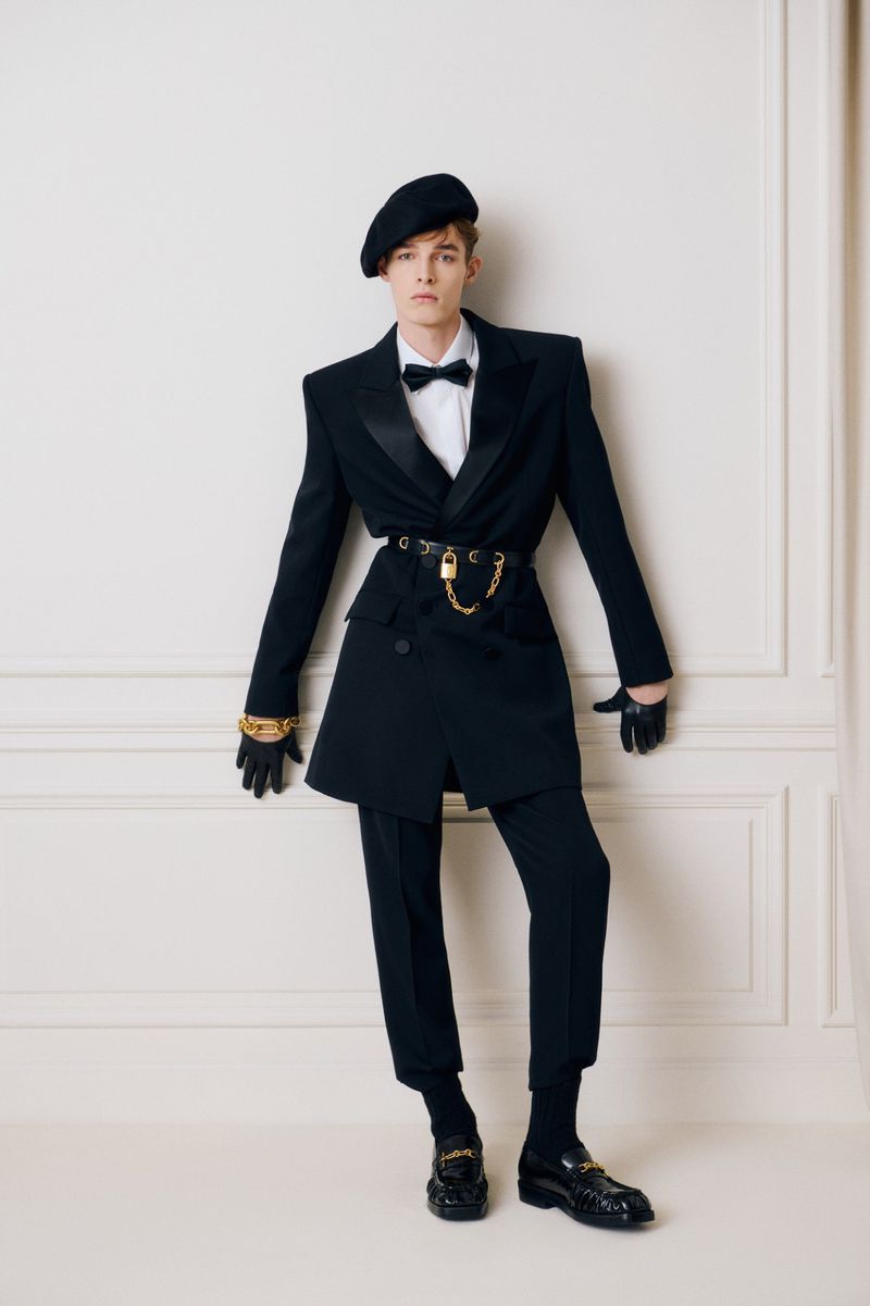 Fashionable person poses in a tailored black suit with a beret, gold chain, and black loafers against a light wall in a studio setting.