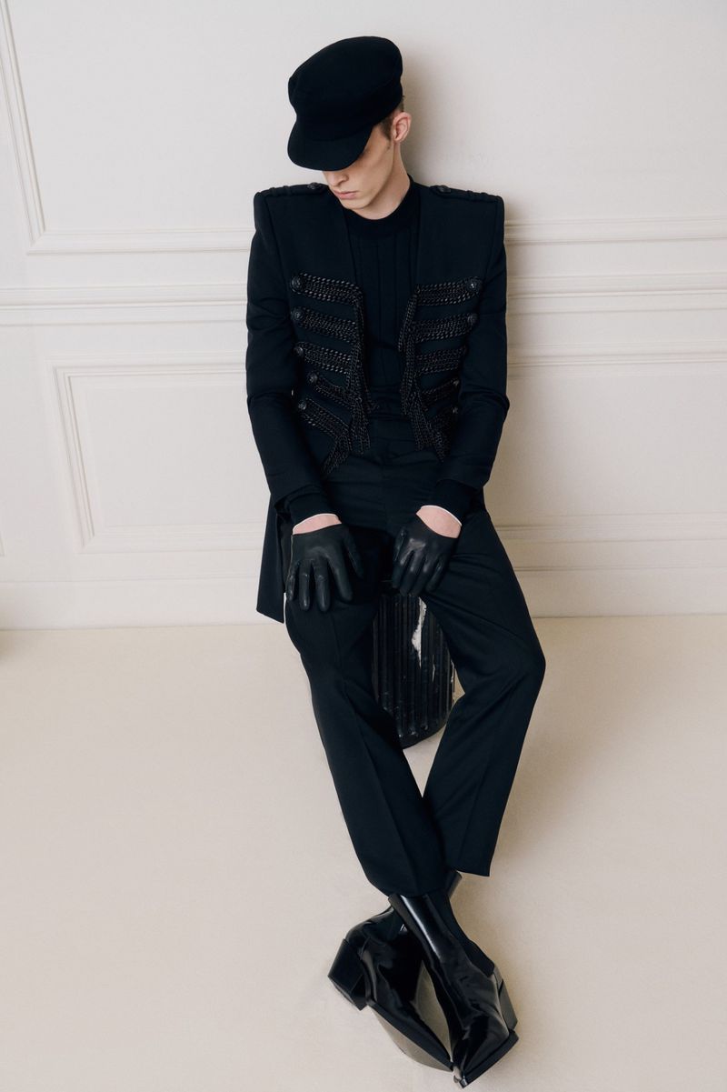 Full-length portrait of a stylish adult male in a black military-style jacket, trousers, cap, and glossy block-heeled boots against a wall.