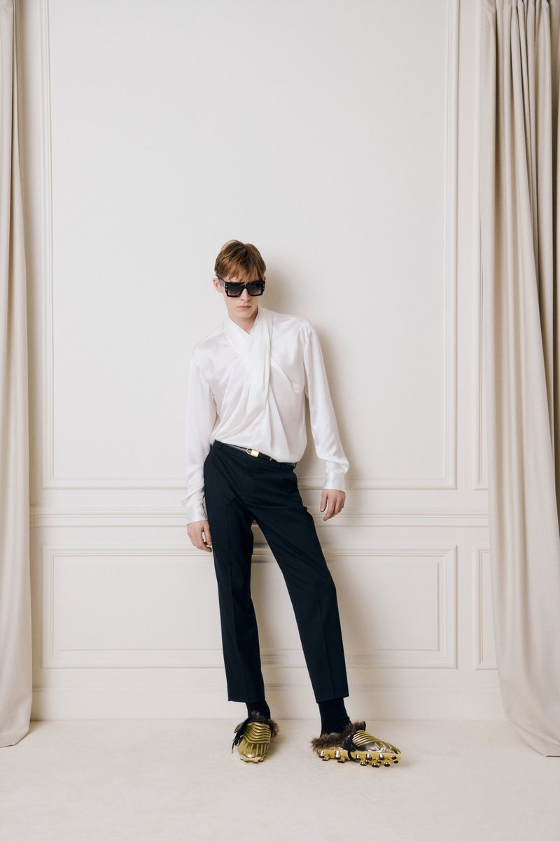 Fashionable adult male posing in a white button-down shirt, dark trousers, and gold shoes against a white studio backdrop.