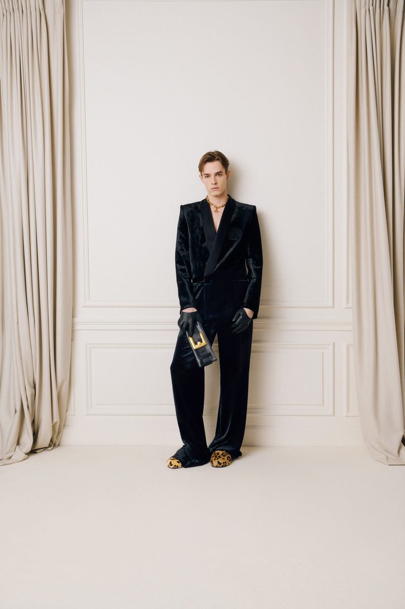 Adult man in velvet suit posing indoors against paneled wall, holding a clutch, wearing black gloves and animal print footwear.