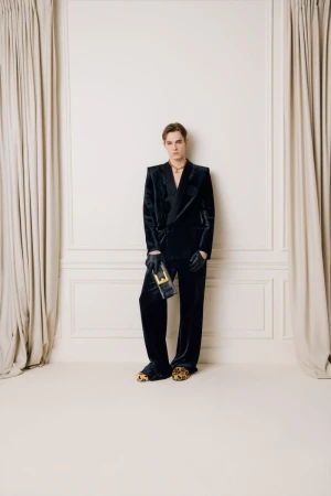 Adult man in velvet suit posing indoors against paneled wall, holding a clutch, wearing black gloves and animal print footwear.