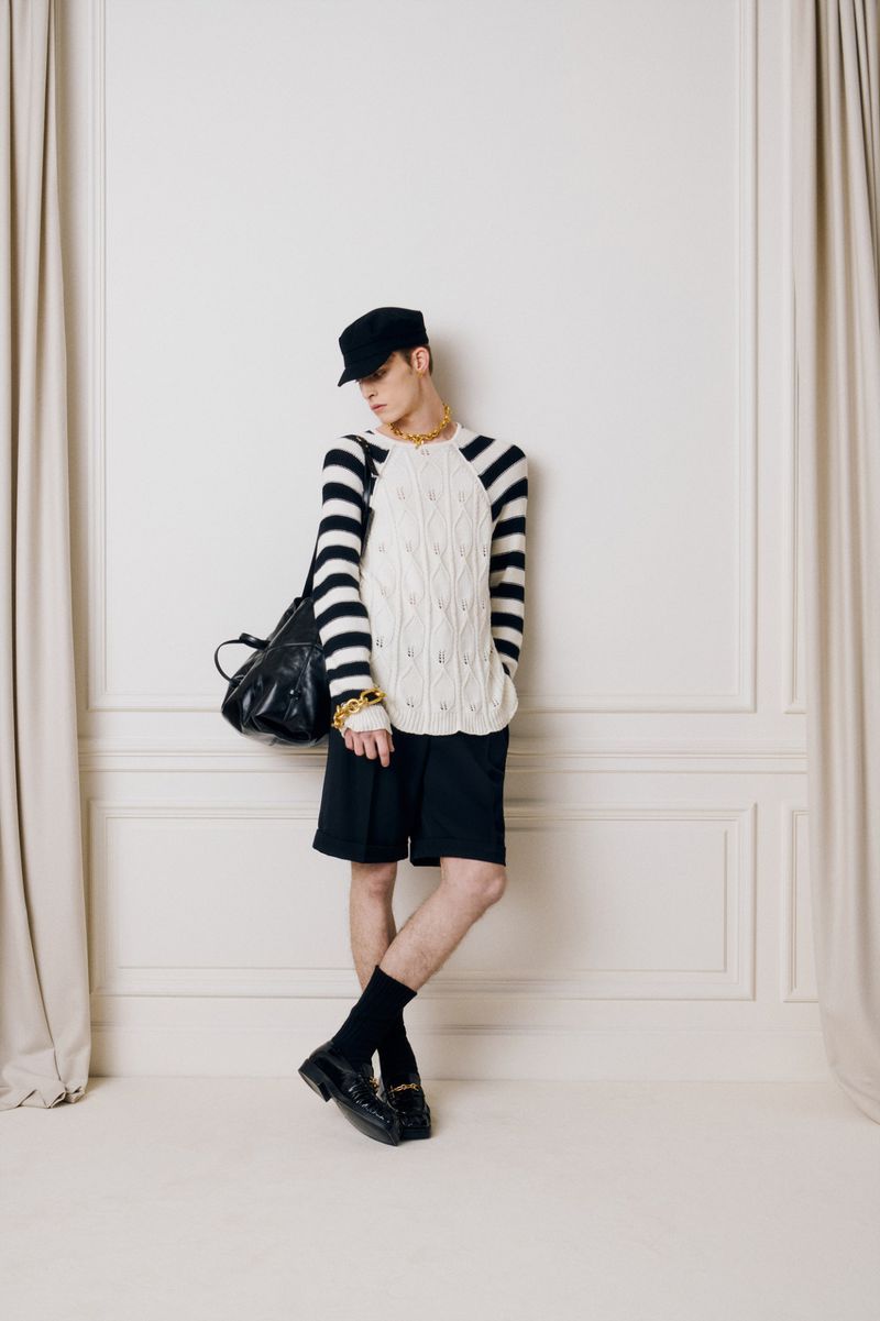 Studio shot of a male model wearing a striped long-sleeve shirt, shorts, cap, gold chain, and loafers, leaning against a white wall.