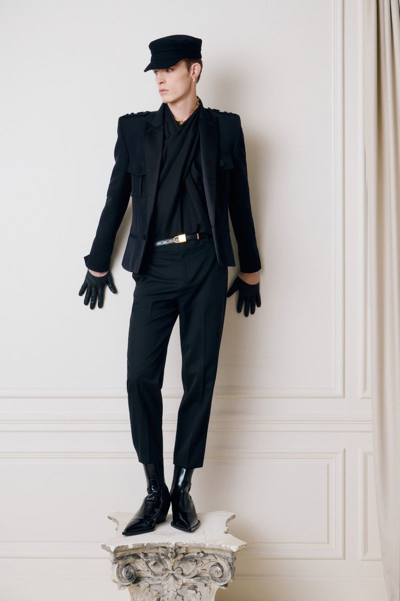 Male fashion model poses in black military-style jacket, pants, cap, and gloves on a pedestal against a neutral wall with draped fabric.