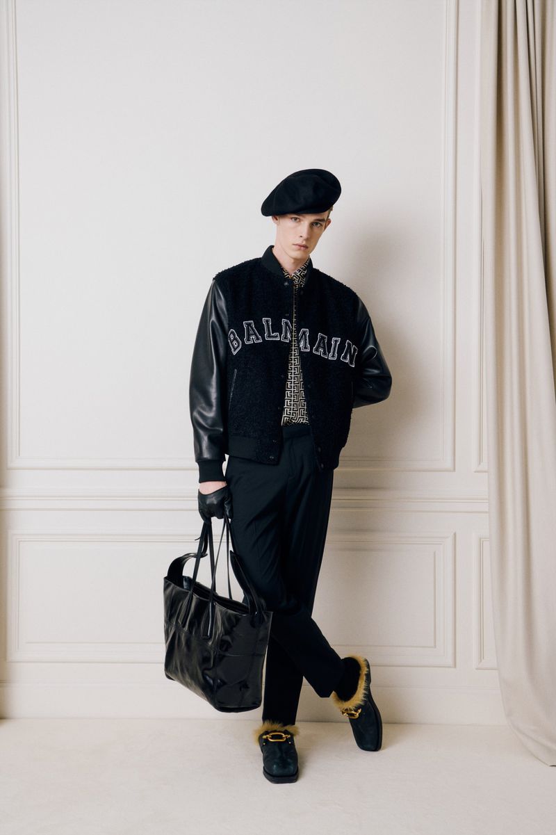 Man in black jacket and beret stands in front of a white wall with a black leather bag. Fashion editorial style.