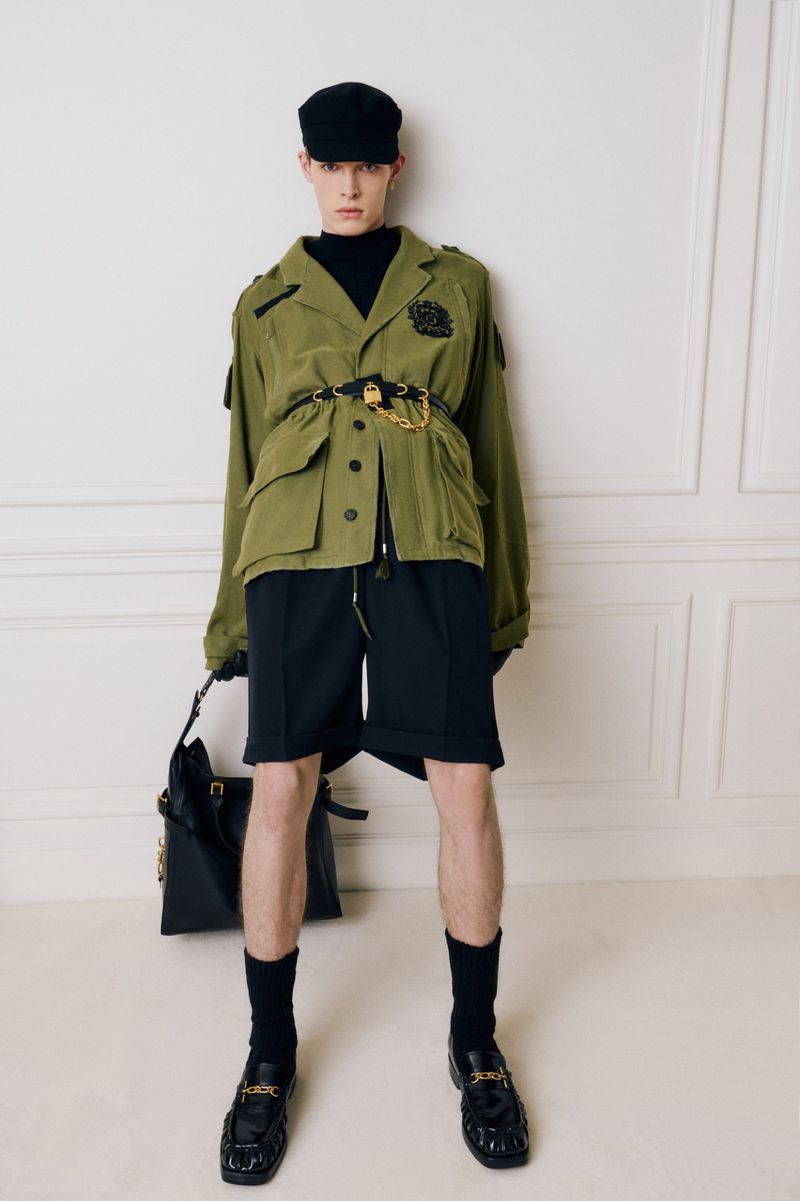 Model in olive green jacket and black shorts holding a leather handbag against a pale panelled wall