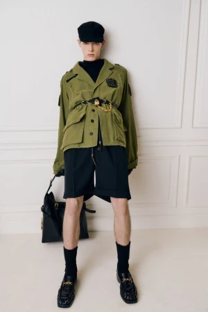 Model in olive green jacket and black shorts holding a leather handbag against a pale panelled wall