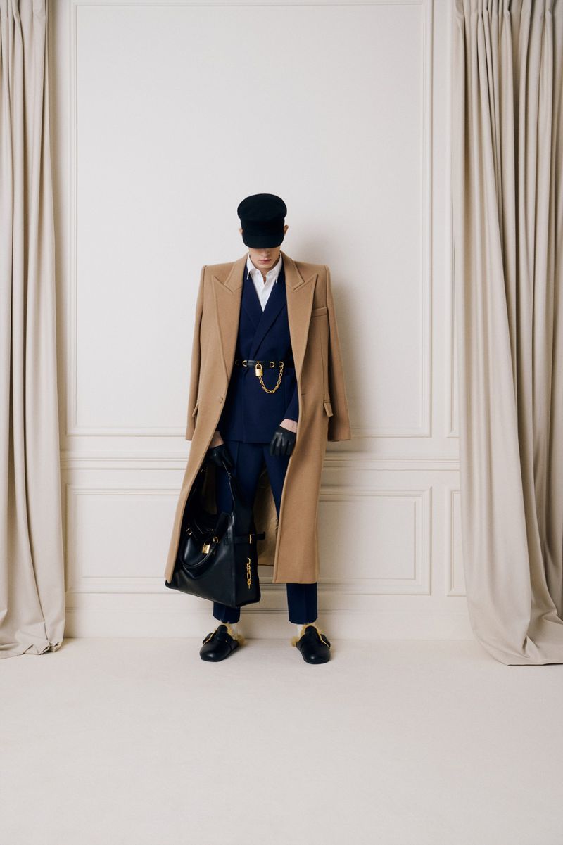 Person wearing a long tan coat, blue suit, black hat, gloves, and loafers with a black leather purse, standing in a photo studio.