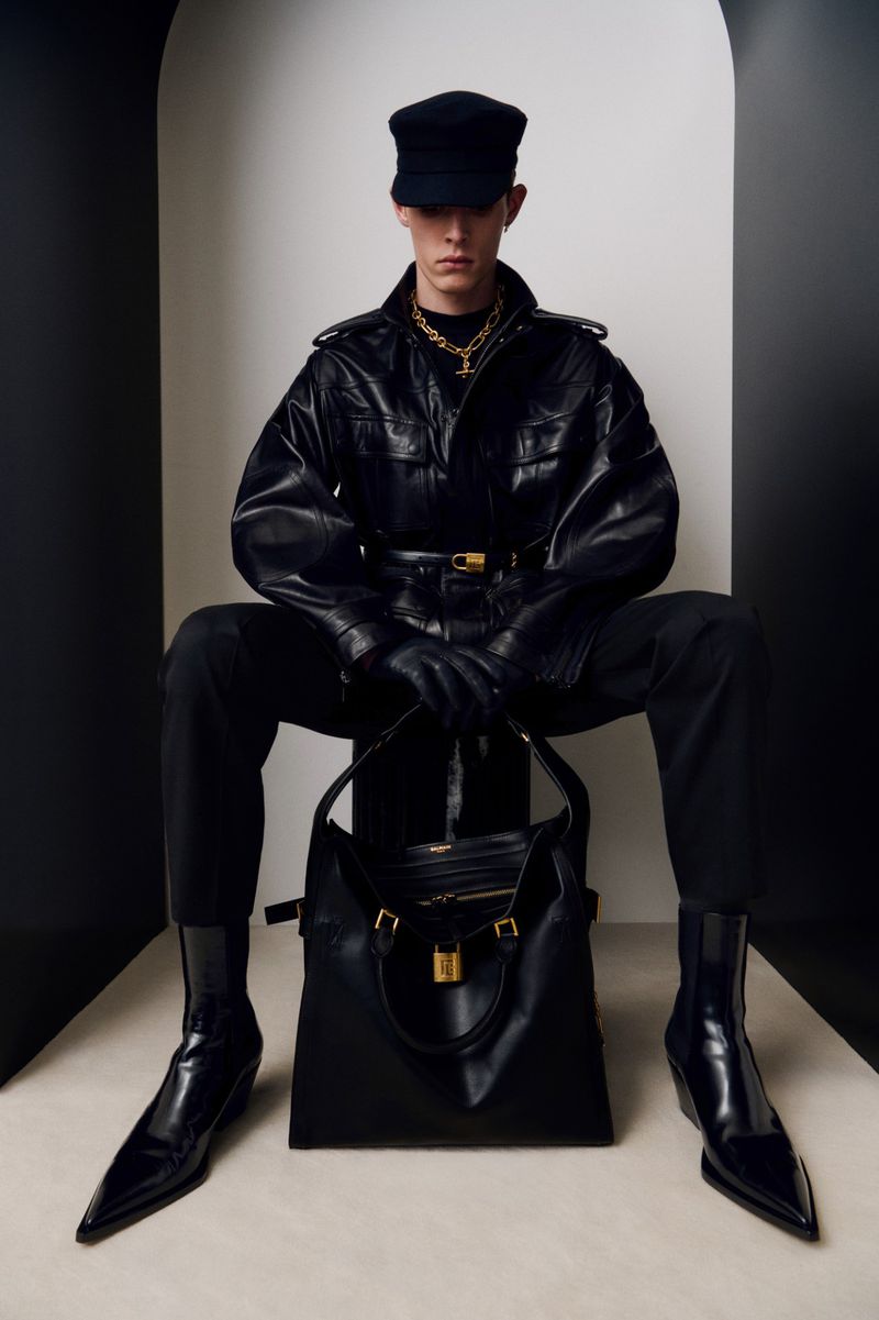 Male fashion model wearing black leather jacket and cap, seated with large black leather handbag in a minimalist studio setting.