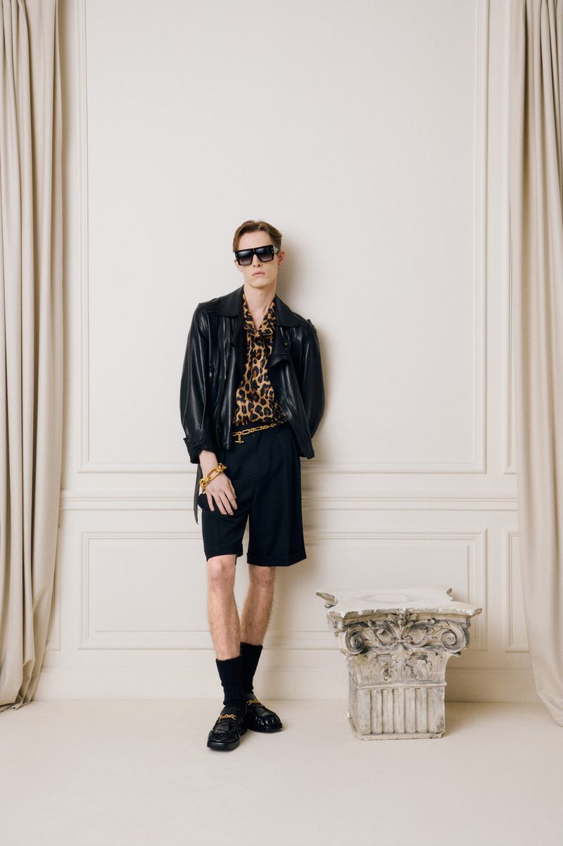 Stylish man in black leather jacket, animal print shirt, and shorts posing against a textured white wall with classic architectural details.