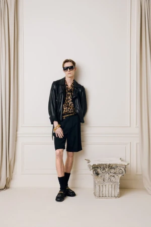 Stylish man in black leather jacket, animal print shirt, and shorts posing against a textured white wall with classic architectural details.