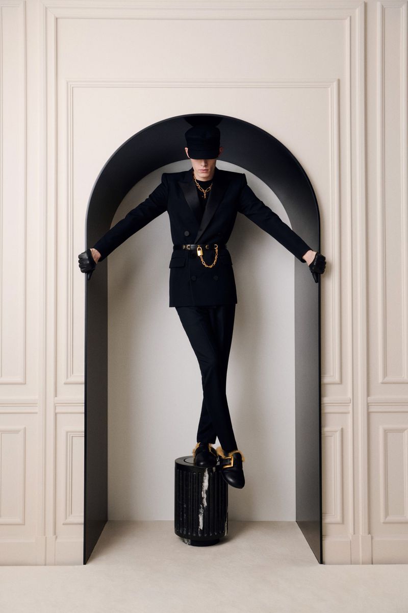 Person in a black suit and cap standing on a cylinder in front of a doorway, arms outstretched, in a light-filled indoor setting.