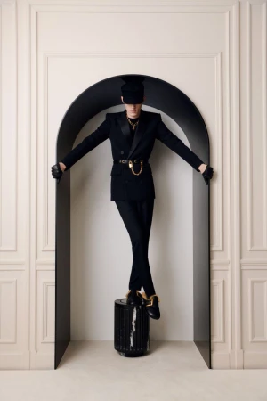 Person in a black suit and cap standing on a cylinder in front of a doorway, arms outstretched, in a light-filled indoor setting.