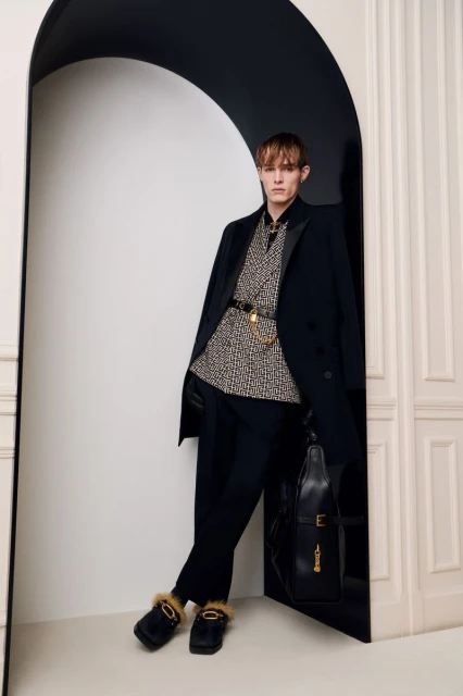 Man in patterned shirt, black coat, and slippers poses in a doorway with a purse. Fashion shot with a neutral color palette.