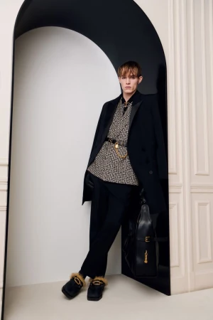 Man in patterned shirt, black coat, and slippers poses in a doorway with a purse. Fashion shot with a neutral color palette.