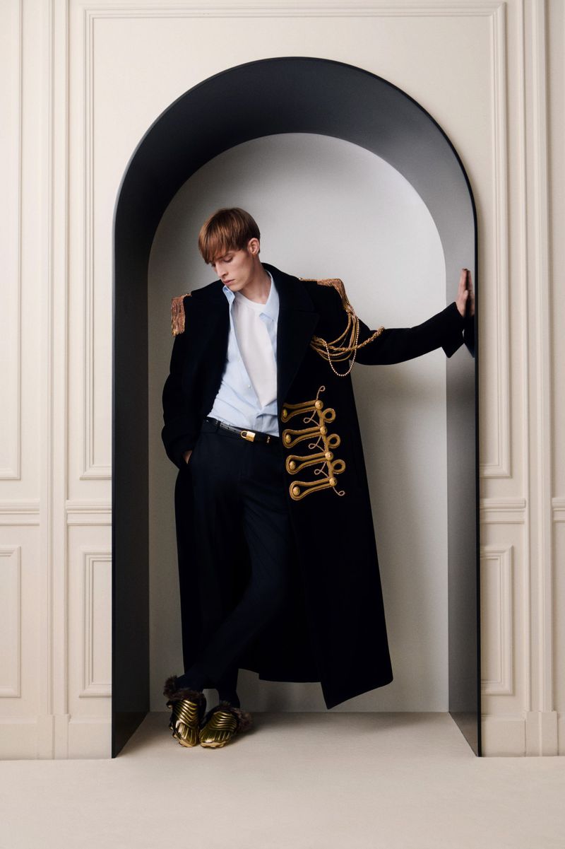 Male fashion model in black coat with gold accents, posing in arched doorway against off-white wall, wearing metallic gold shoes.