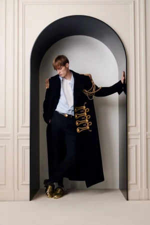 Male fashion model in black coat with gold accents, posing in arched doorway against off-white wall, wearing metallic gold shoes.