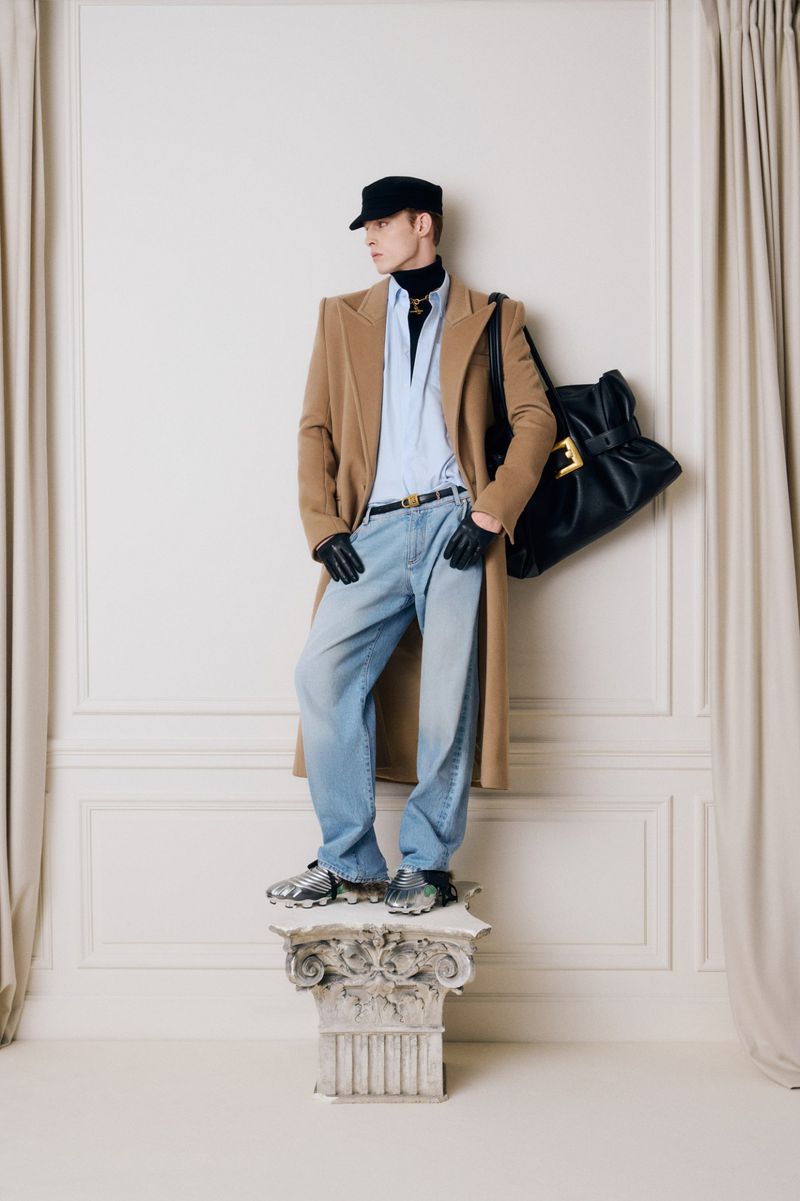 A male fashion model poses in a tan overcoat, blue jeans, and black accessories on a decorative plinth in a studio setting.