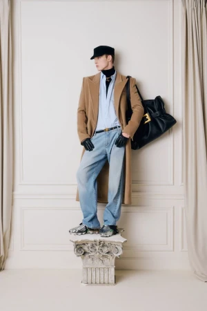 A male fashion model poses in a tan overcoat, blue jeans, and black accessories on a decorative plinth in a studio setting.