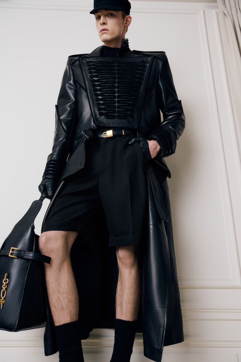 Model wearing a structured black leather coat, matching shorts, cap, gloves, and holding a large leather bag indoors.