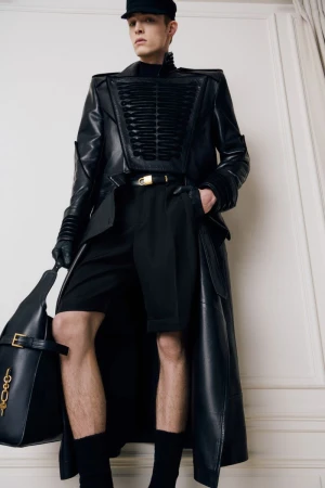 Model wearing a structured black leather coat, matching shorts, cap, gloves, and holding a large leather bag indoors.