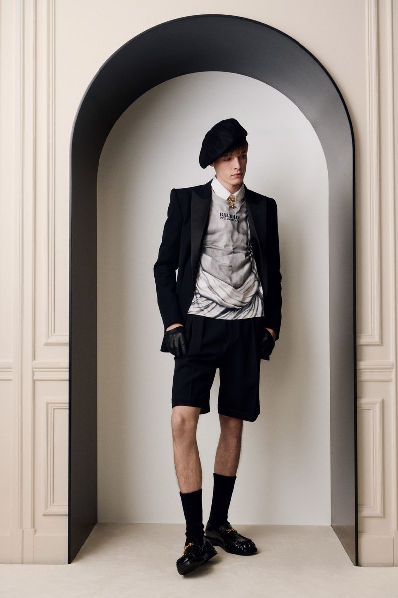 Model wearing a black beret, blazer, shorts, and graphic top standing within an arched architectural opening.