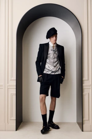 Model wearing a black beret, blazer, shorts, and graphic top standing within an arched architectural opening.