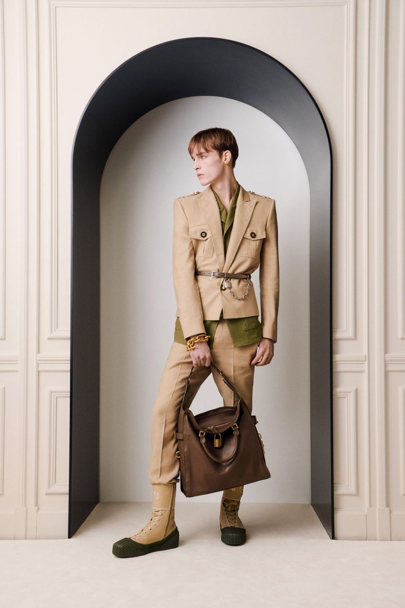 Male fashion model in neutral suit with brown leather tote bag poses in front of an arched wall design in studio setting.