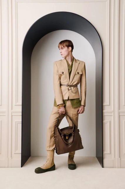 Male fashion model in neutral suit with brown leather tote bag poses in front of an arched wall design in studio setting.