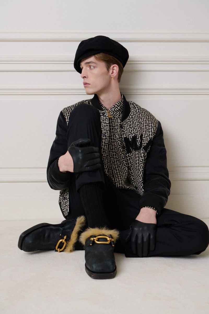 Adult male wearing a black beret, patterned jacket, and black boots with fur trim, seated against a neutral wall. Fashion editorial.