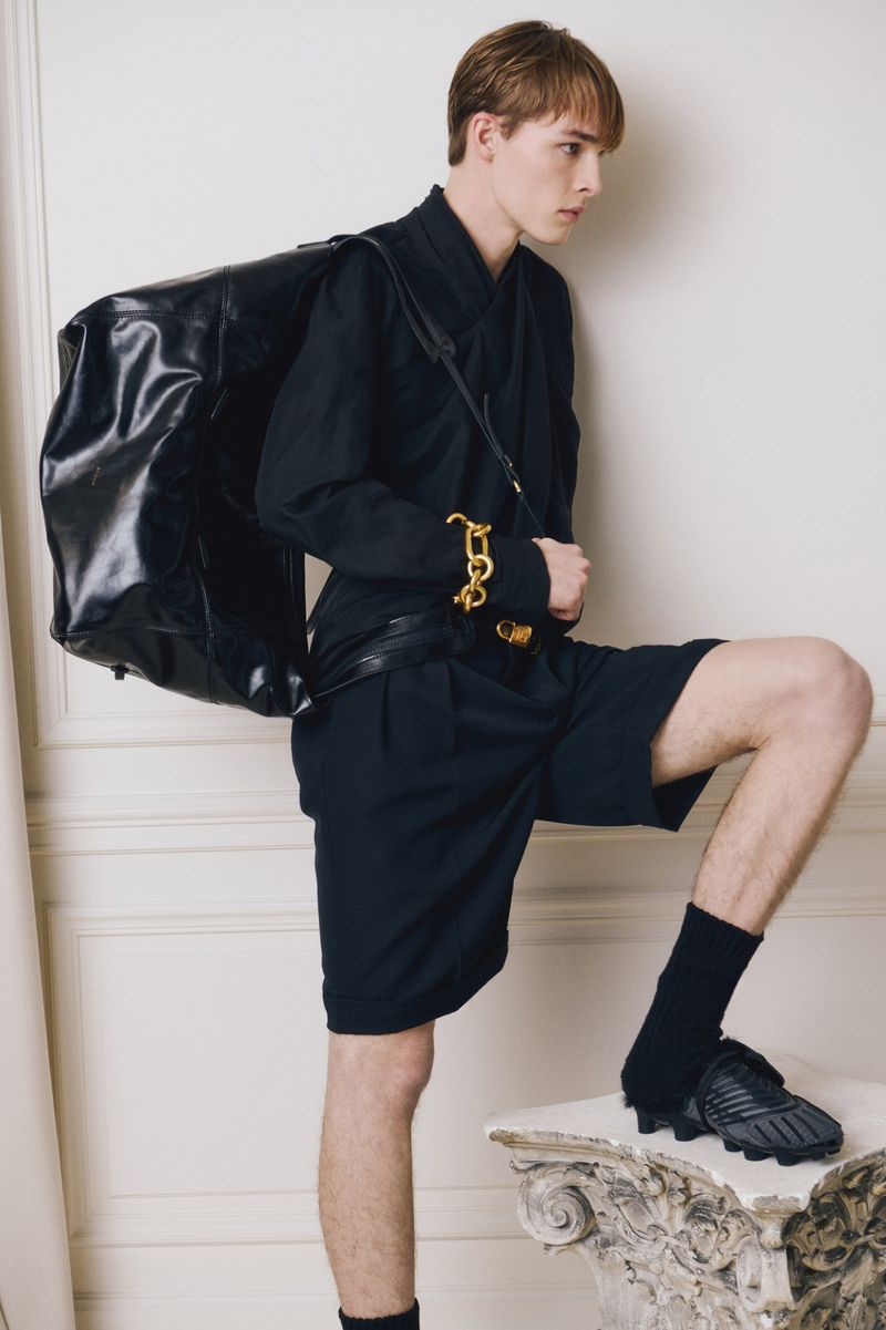 A fair-skinned young adult man poses indoors with a black leather duffel bag, wearing a black shirt, shorts, socks, and dark shoes.