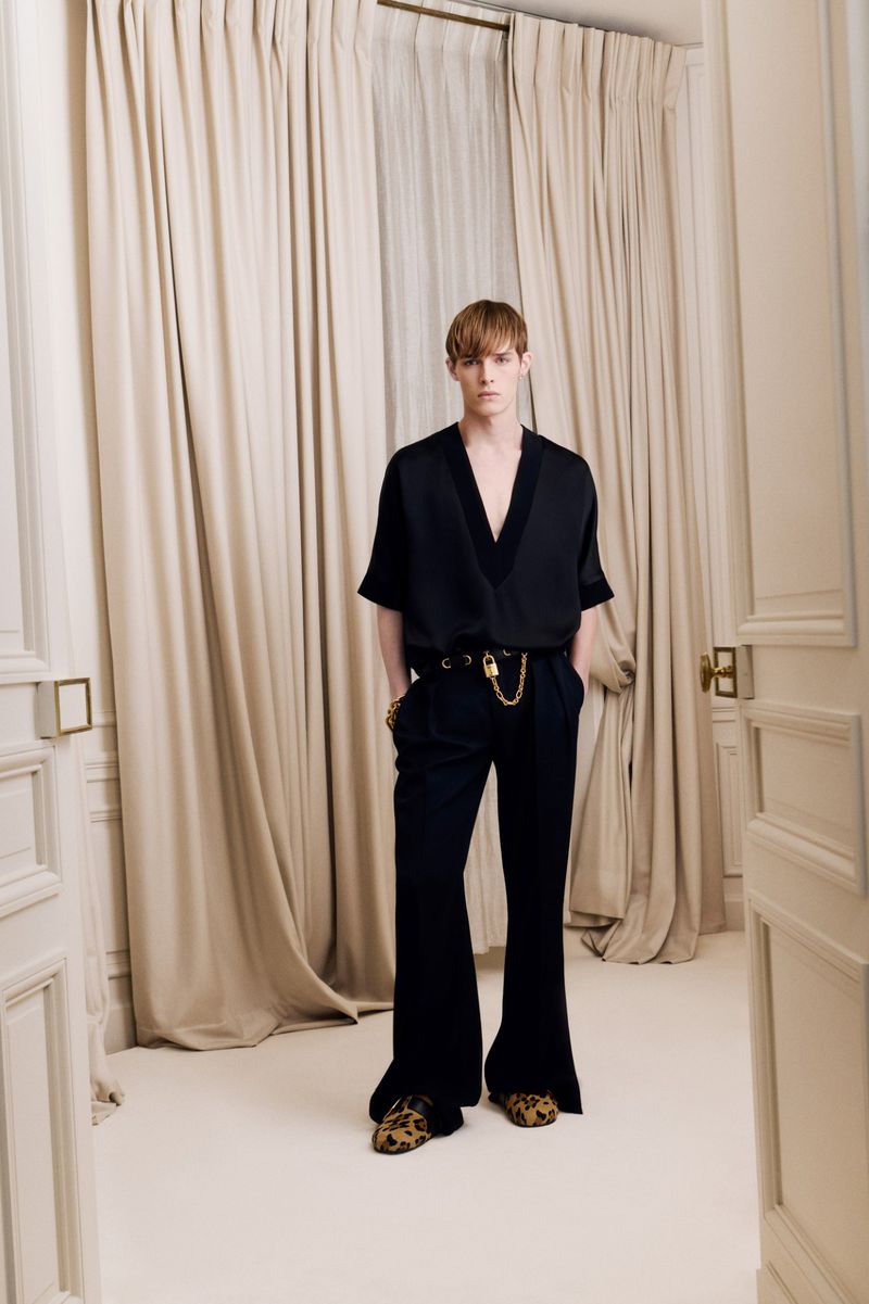 Adult male fashion model with auburn hair wearing a black blouse and black flared pants poses in front of a white curtain backdrop.