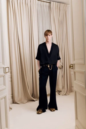 Adult male fashion model with auburn hair wearing a black blouse and black flared pants poses in front of a white curtain backdrop.