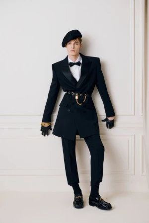 Model wearing a black suit jacket, beret, and bow tie standing against a wall with paneling details for Balmain Pre-Fall 2025 lookbook.