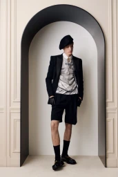 Model wearing a black beret, blazer, shorts, and graphic top standing within an arched architectural opening.