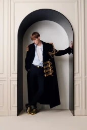 Model in black military-style coat with gold braiding leaning inside an architectural arched alcove