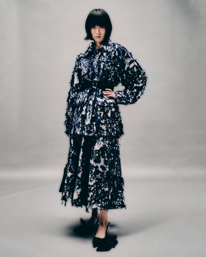 A full-shot of an adult woman posing in a studio setting, wearing a black-and-white patterned, two-piece outfit and black high heels.