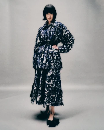 A full-shot of an adult woman posing in a studio setting, wearing a black-and-white patterned, two-piece outfit and black high heels.