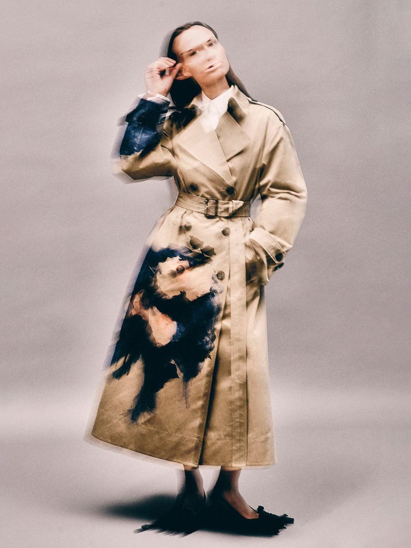 A woman in a tan trench coat poses against a light gray background, holding up the frame of her glasses with her right hand.