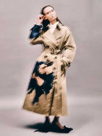 A woman in a tan trench coat poses against a light gray background, holding up the frame of her glasses with her right hand.