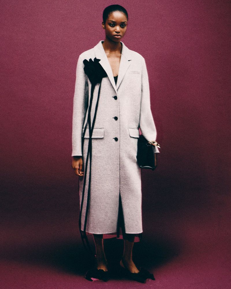 Fashion editorial shot of a Black female model wearing a long grey wool coat with black floral design against a plum studio backdrop.