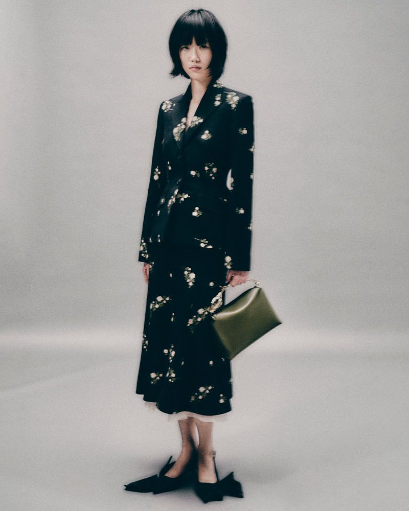 A stylish Asian woman with short hair poses in a patterned coat dress with an olive-green handbag against a neutral background in studio lighting.