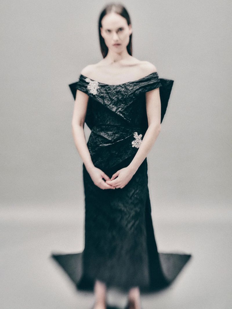 A woman in a black off-the-shoulder gown stands in front of a grey background, creating an editorial fashion image with an elegant aesthetic.
