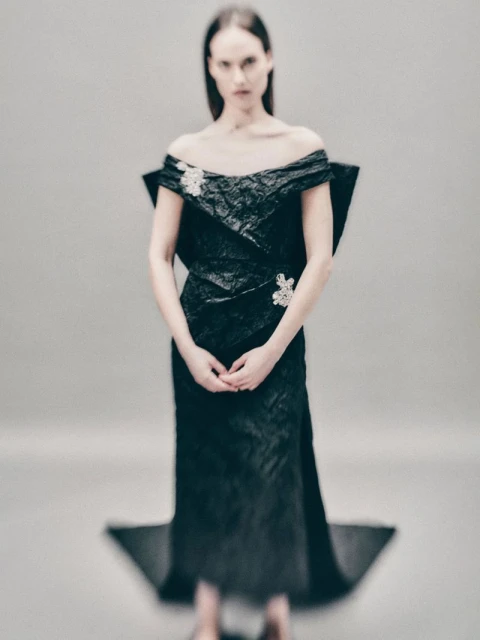 A woman in a black off-the-shoulder gown stands in front of a grey background, creating an editorial fashion image with an elegant aesthetic.